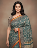A woman modeling a teal blue and beige patterned saree with a gold and orange border, set against a solid brown studio background.